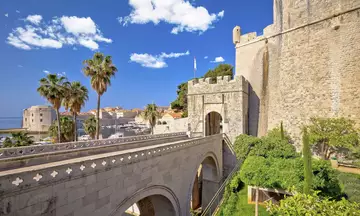 Entrada principal a las murallas de Dubrovnik, con puente de piedra, palmeras y vistas al puerto y a la ciudad amurallada.