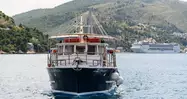El barco Arupinum navegando frente a la costa de Dubrovnik con un crucero y puente al fondo.
