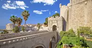 Entrada principal a las murallas de Dubrovnik, con puente de piedra, palmeras y vistas al puerto y a la ciudad amurallada.