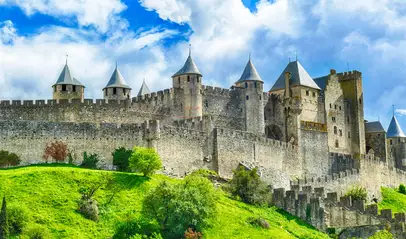 Vista de las murallas y torres medievales de Carcasona entre colinas verdes, bajo un cielo dramático.