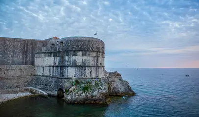 Murallas medievales de Dubrovnik al atardecer, con torre fortificada sobre el mar Adriático y embarcaciones pequeñas en el horizonte.