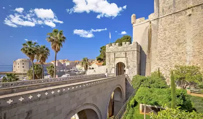 Entrada principal a las murallas de Dubrovnik, con puente de piedra, palmeras y vistas al puerto y a la ciudad amurallada.