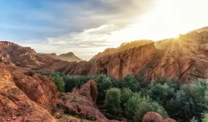 Paisaje del Alto Atlas al atardecer con formaciones rocosas rojizas y un oasis de vegetación frondosa entre las montañas.